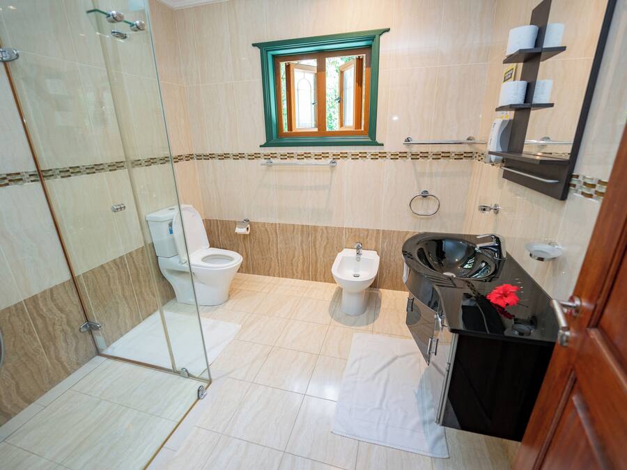 A bathroom with a glass shower enclosure, two toilets, a bidet, a vanity with a mirror, and a window with a view.