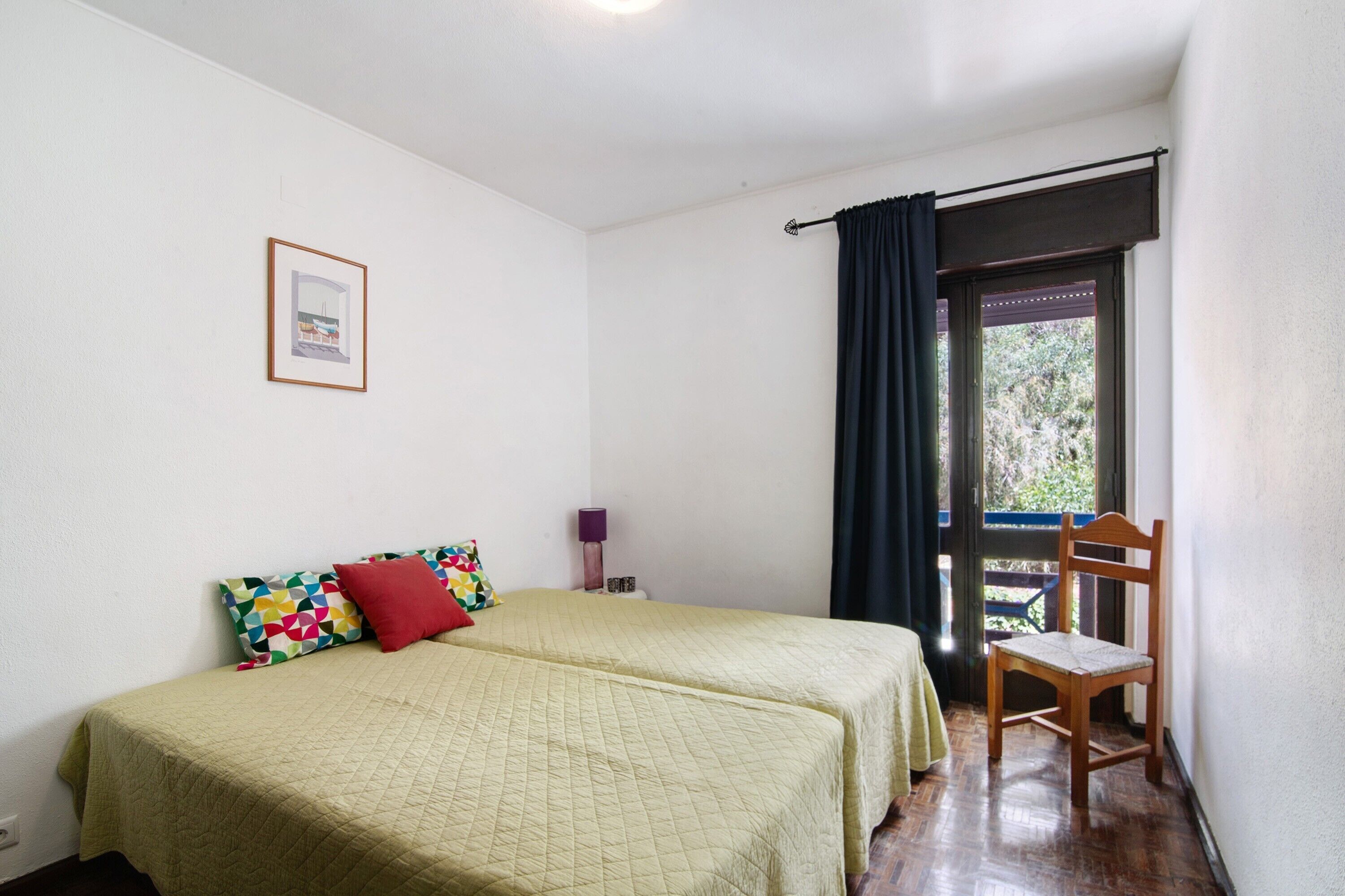 A room with two beds, a chair, a window with curtains, a framed picture on the wall, and a door leading outside.