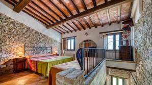 A rustic room with stone walls, a wooden ceiling, a bed with a green bedspread, a small table with a lamp, and a staircase. - Farmhouse 'Casale Storico Xvii Sec - Tulipano' with Mountain View and Shared Pool (Giano dell'Umbria)