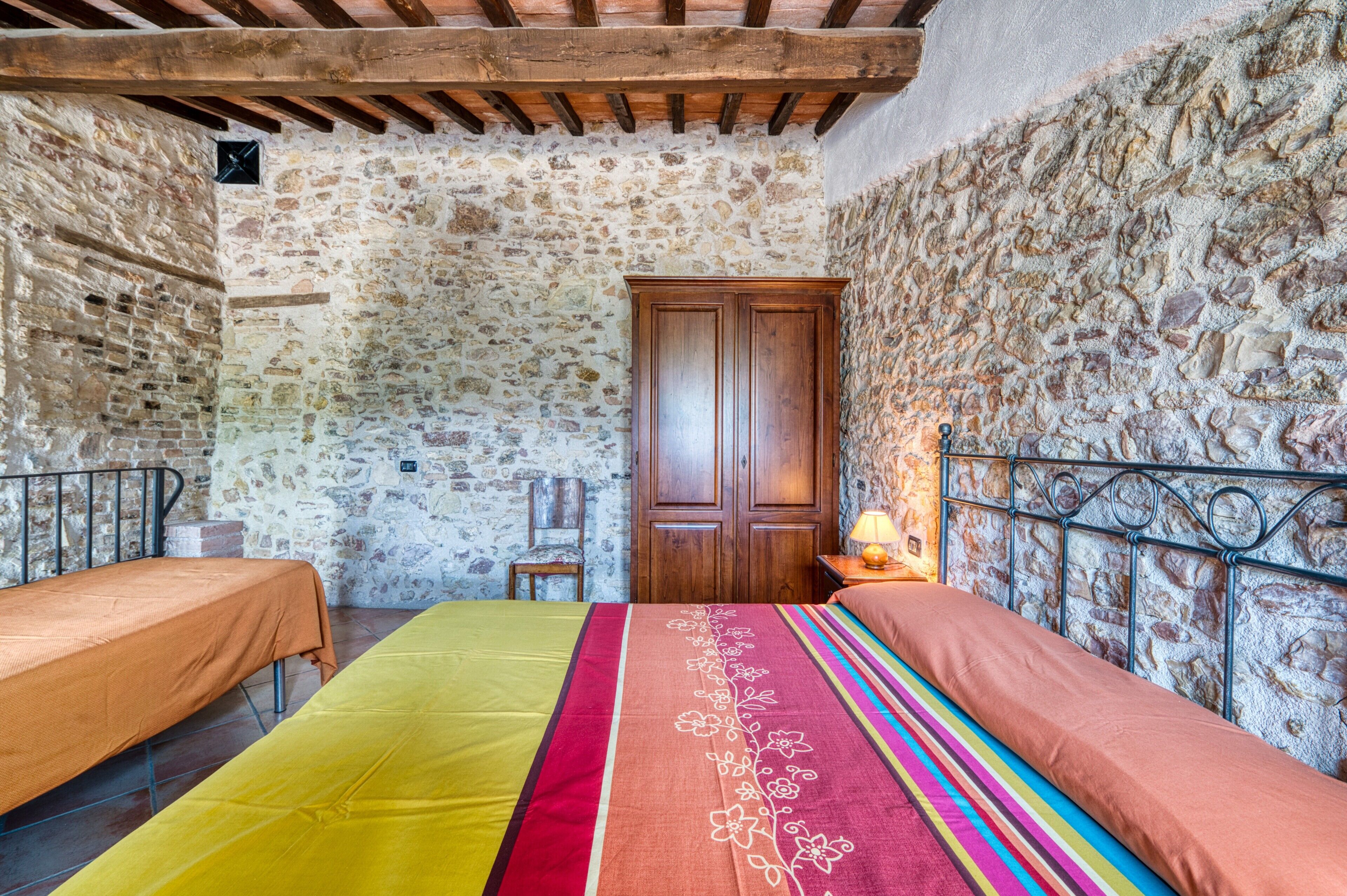 A room with stone walls, a bed with a colorful bedspread, a wooden wardrobe, and a metal headboard.