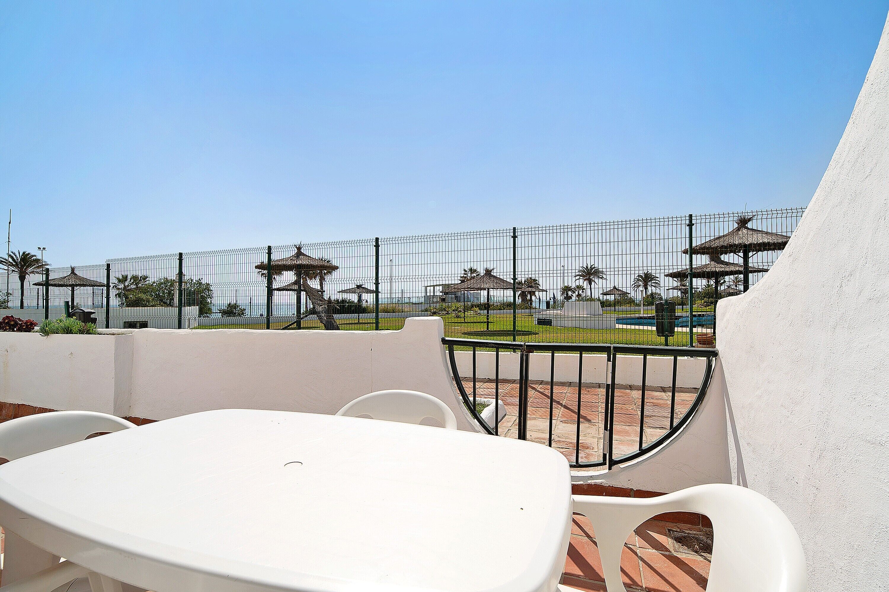 A terrace with a white table and chairs, a metal fence, and a view of a park with umbrellas.
