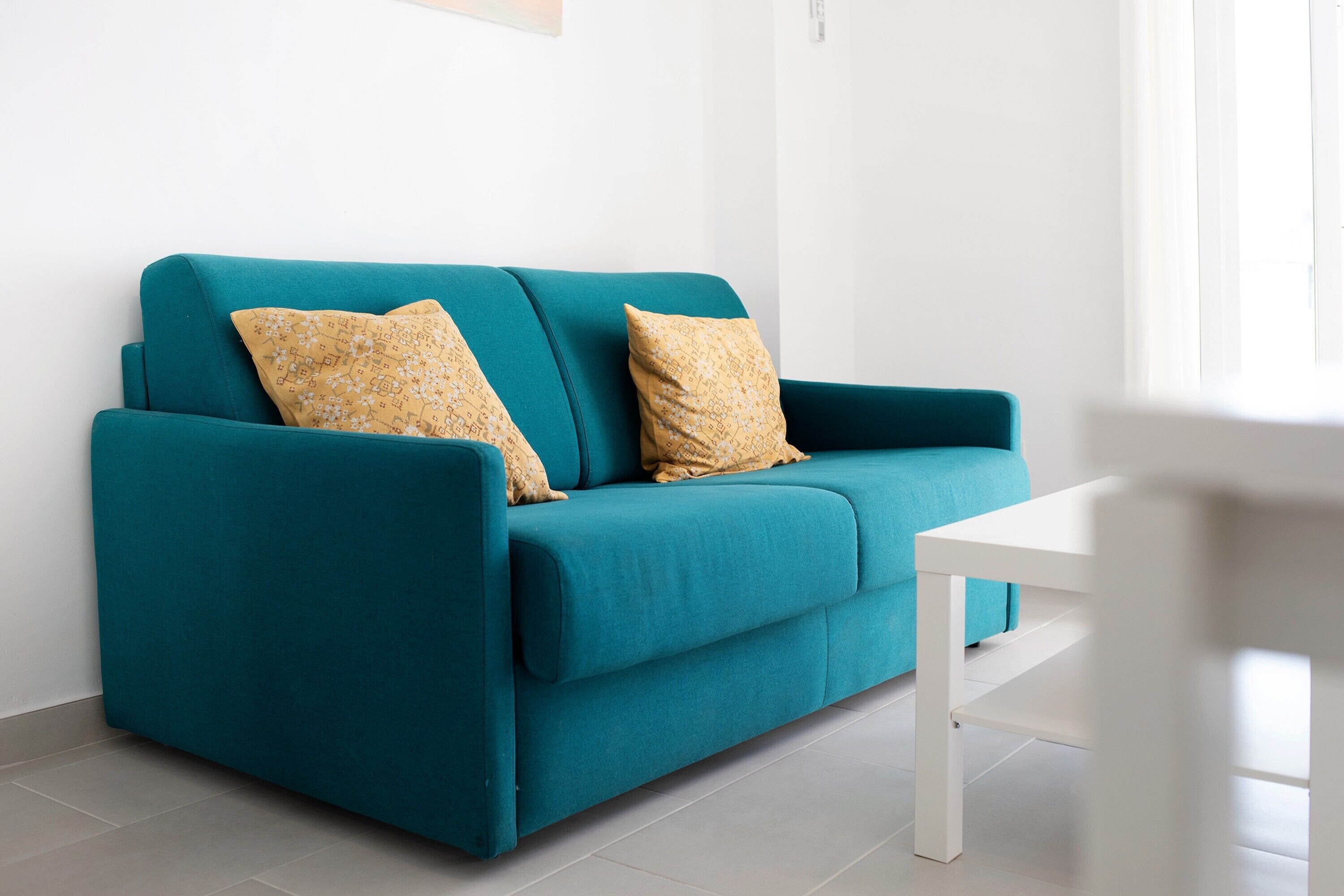 A teal sofa with patterned cushions, a white table, and a plain white wall.
