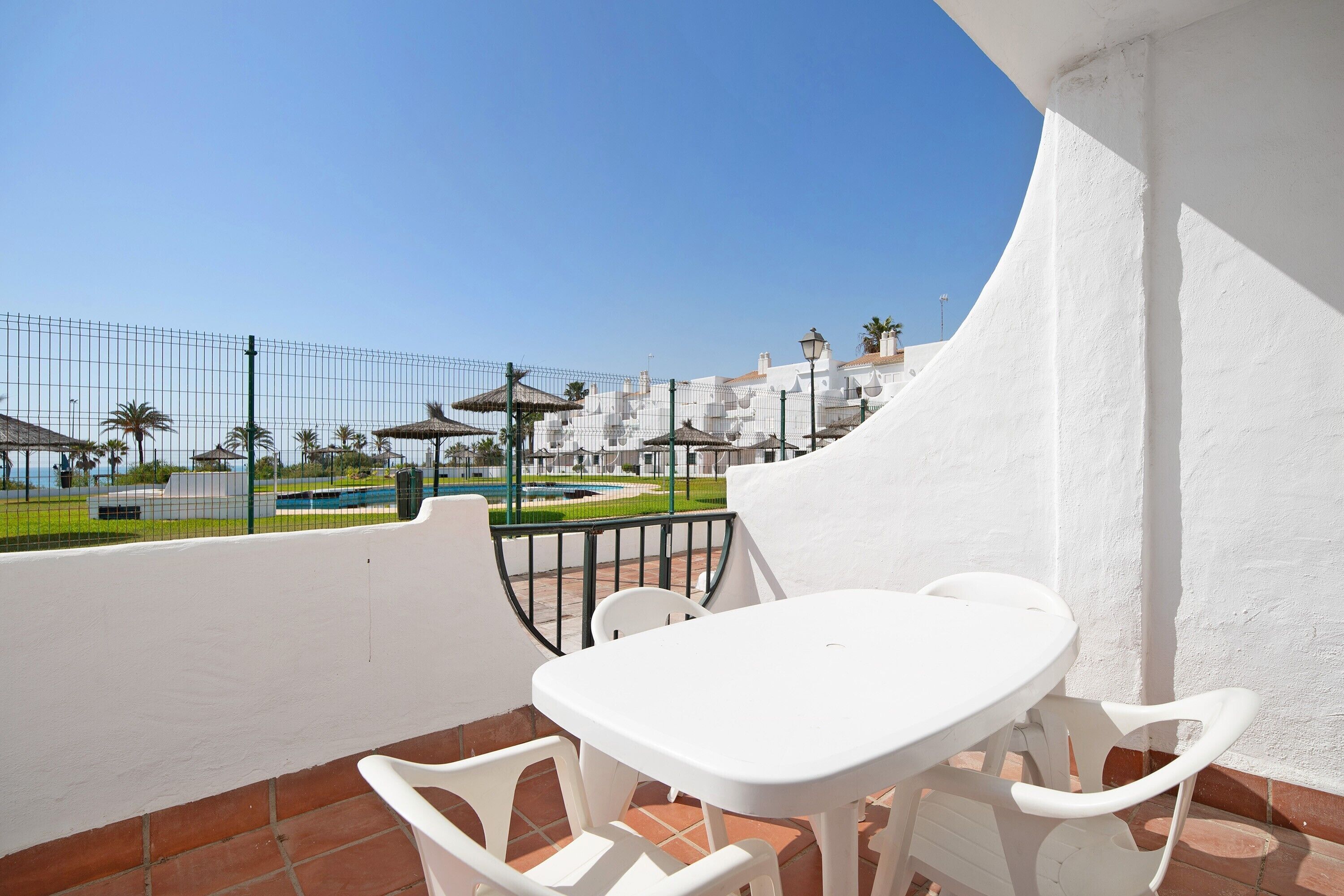 Varanda com mesa e cadeiras brancas, vista para uma piscina e vegetação, e um edifício branco ao fundo.