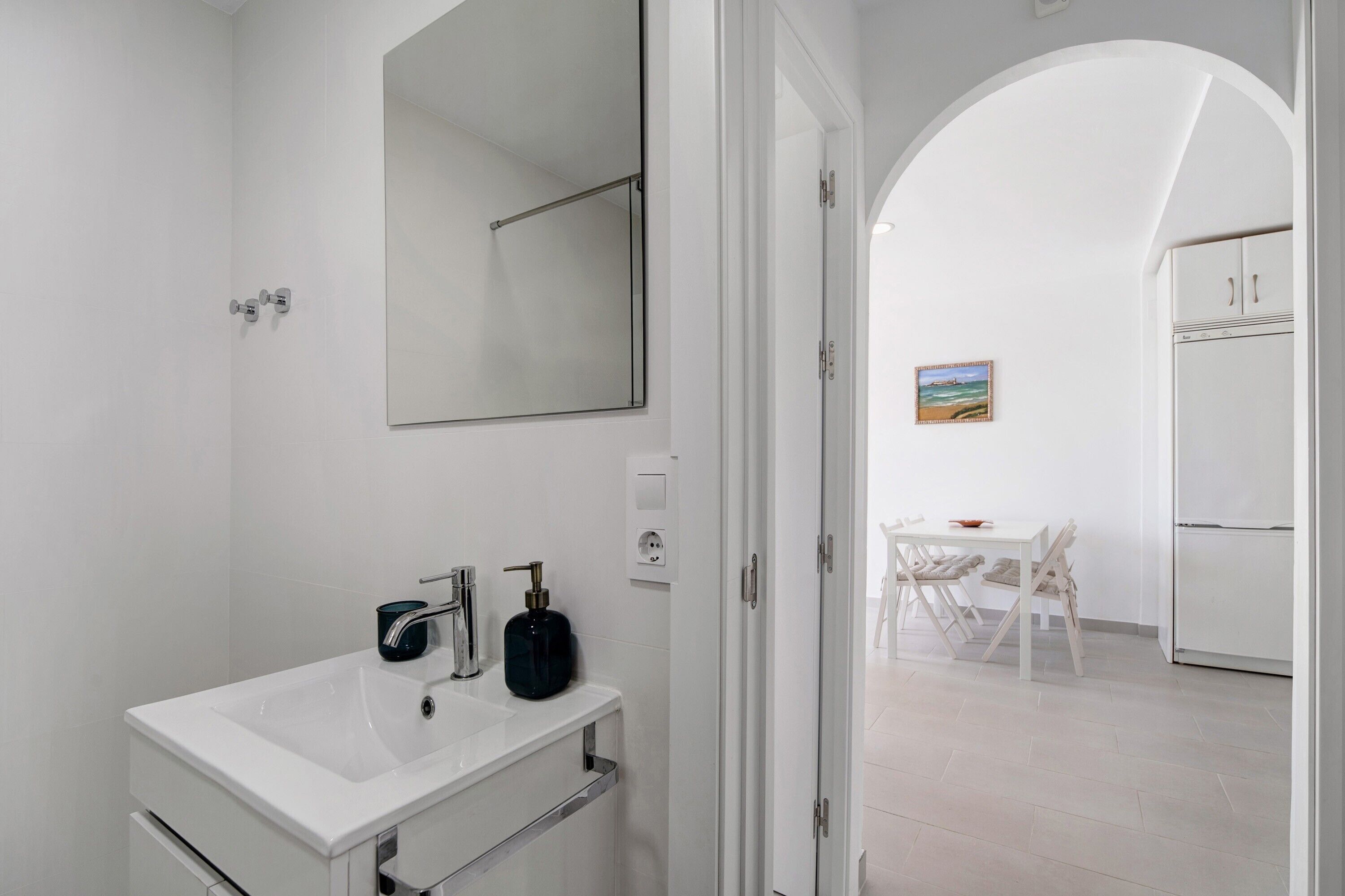 Un baño moderno con lavamanos, espejo y ducha blancos. Se ve un comedor con una mesa y sillas a través de un arco.