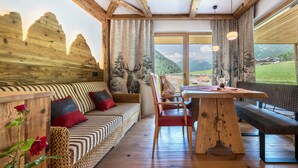 Ein Holzinnenraum mit einer Couch, einer Bank und einem Esstisch, große Fenster mit Blick auf die Berge.