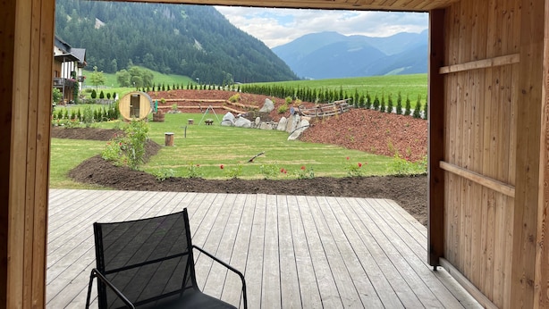 Eine Holzterrasse mit einem Stuhl, Blick auf eine gebirgige Landschaft und ein Gebäude mit Balkon.