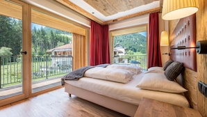 Ein Schlafzimmer mit einem Bett, einem hölzernen Kopfteil, einem Nachttisch und großen Fenstern mit Blick auf ein Haus und Bäume.