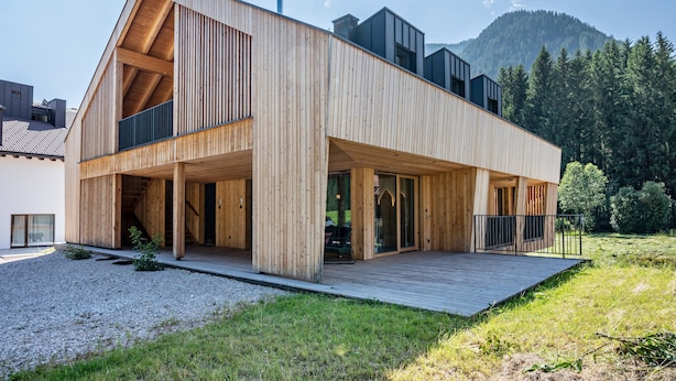 Une maison moderne en bois, dotée d’une terrasse, de grandes fenêtres et offrant une vue sur les montagnes.
