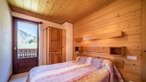 Une pièce aux murs lambrissés, avec un lit, une fenêtre donnant sur une montagne et une porte munie d’un rideau.