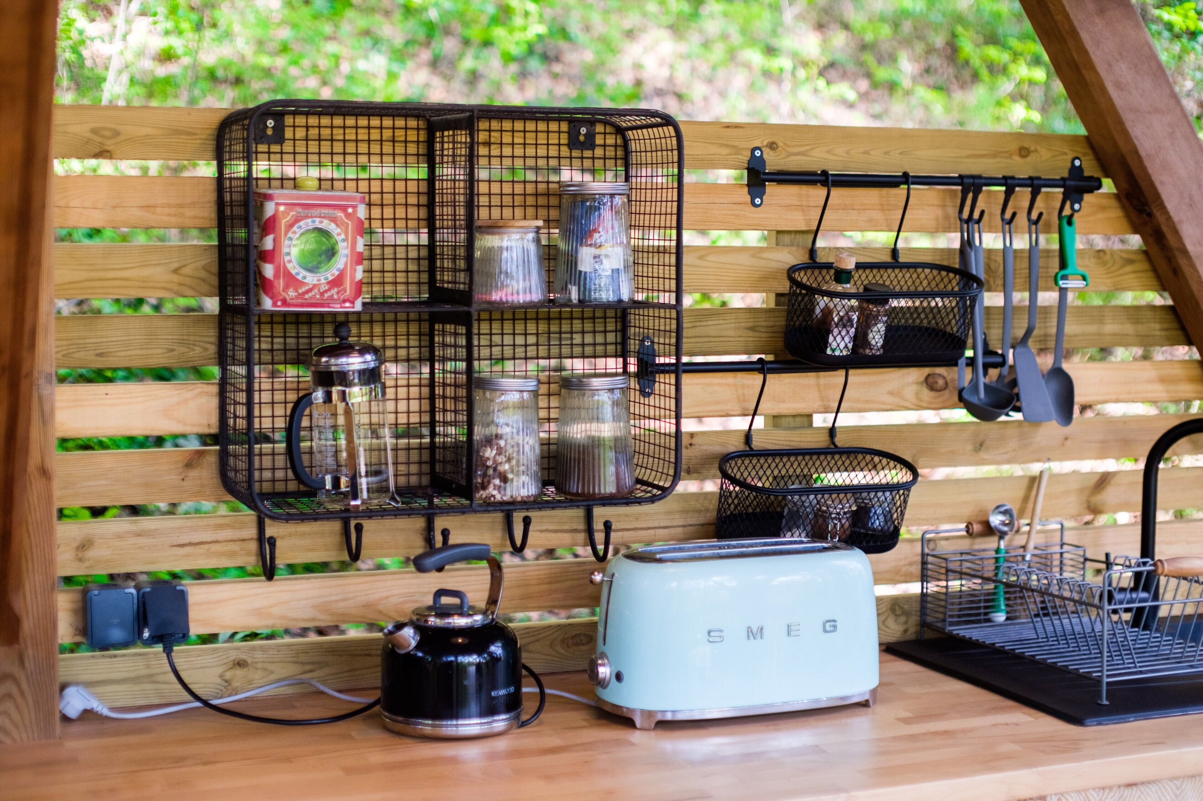 Een keukenhoek met een houten aanrecht, een retro Smeg-toaster, een waterkoker en een rek met potten en keukengerei.
