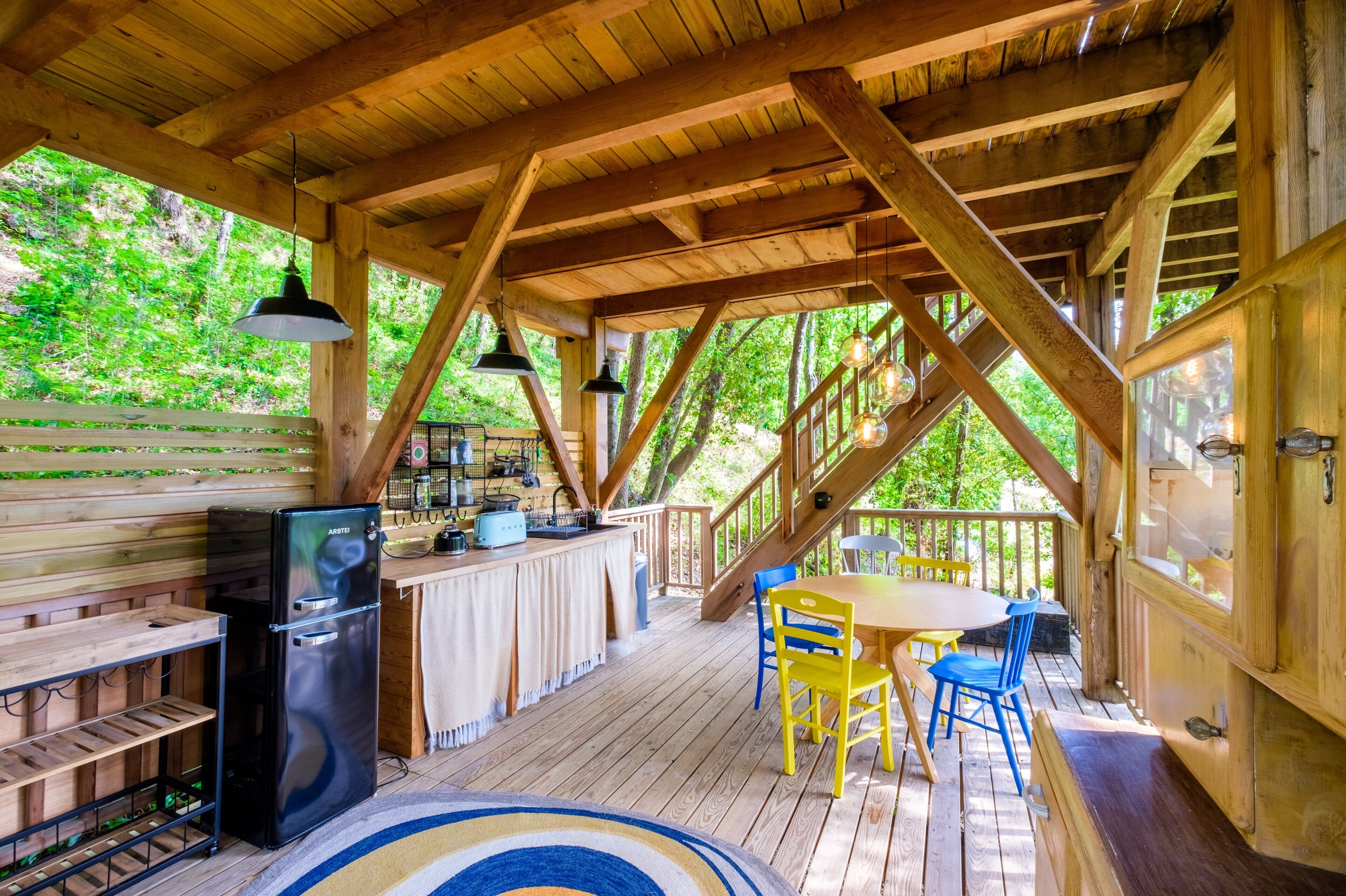 Eine Holzterrasse mit Esstisch, Stühlen, einem Kühlschrank und hängenden Pendelleuchten.