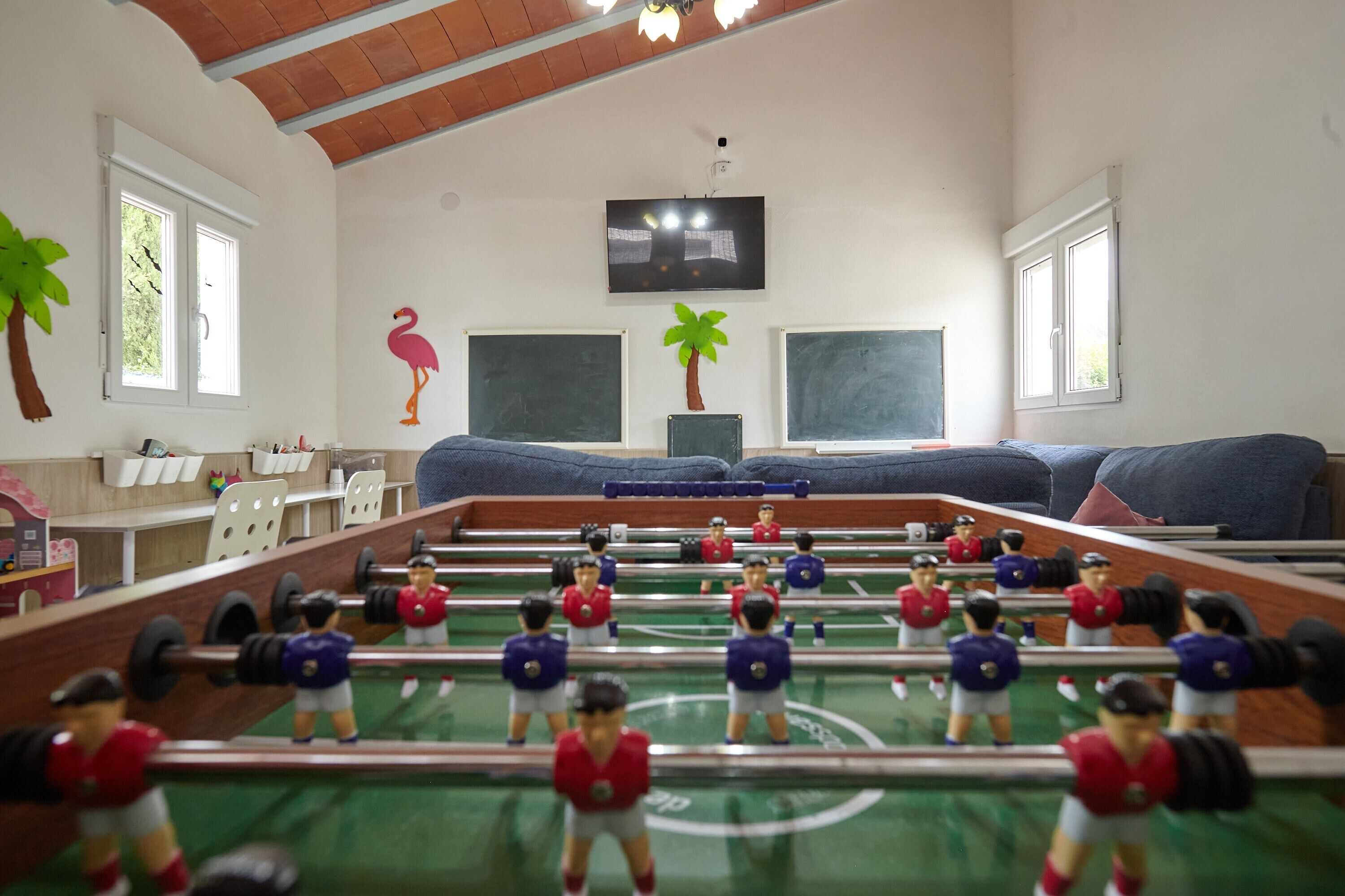 A room with a foosball table, a TV mounted on the wall, and two windows with white frames.
