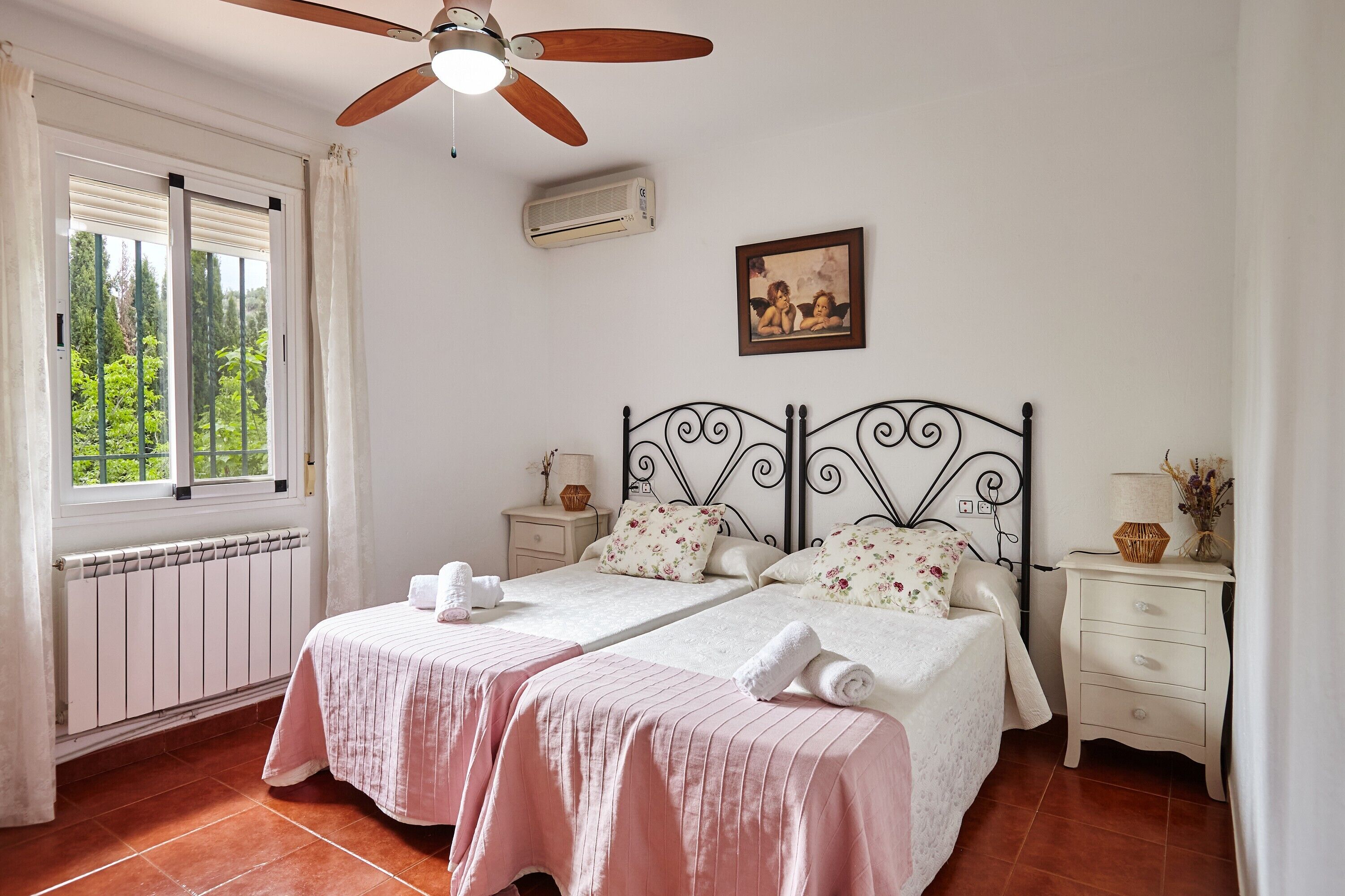 Un dormitorio con dos camas, ventilador de techo, ventana con cortinas, radiador, mesita de noche con lámpara y un cuadro en la pared.