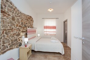 A bedroom with a stone wall, a bed with white bedding, a nightstand with a lamp, and a window with a curtain.
