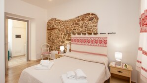 Une chambre d’hôtel avec un mur en pierre, un lit avec des serviettes soigneusement pliées, une chaise, une table de chevet avec une lampe et un petit meuble en bois.