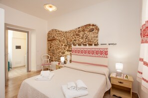 A hotel room with a stone wall, a bed with neatly folded towels, a chair, a bedside table with a lamp, and a small wooden cabinet.