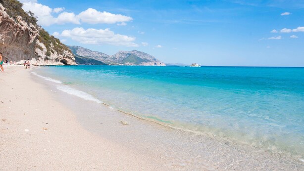 Une plage bordée d’une eau turquoise et limpide, une falaise rocheuse et un bateau aperçu au loin.