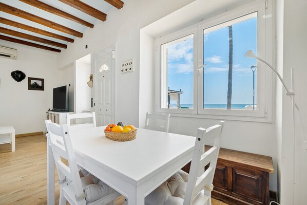 Una mesa de comedor blanca con sillas, una cesta de fruta y vistas al mar a través de una ventana.