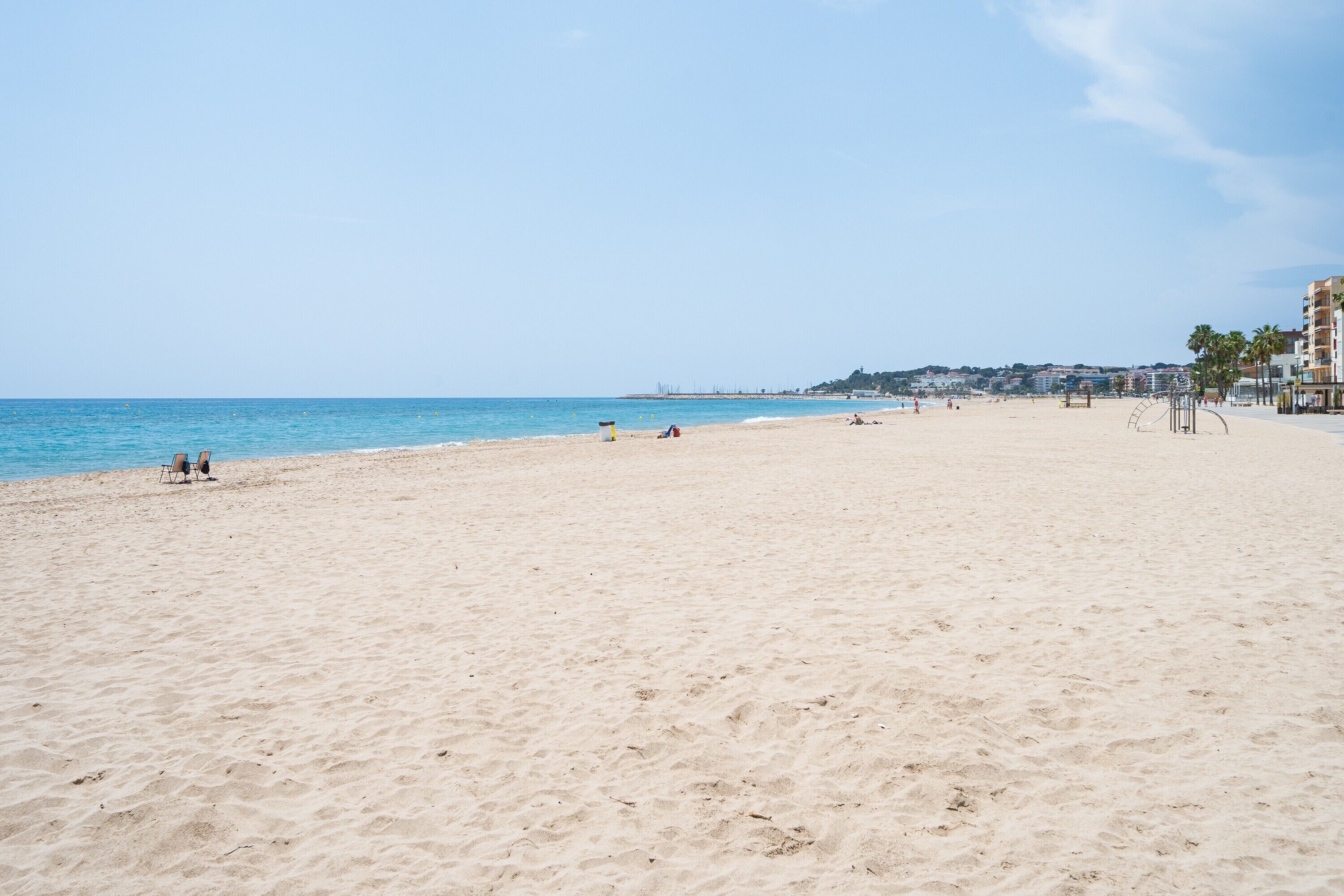 A wide sandy beach with a clear blue sky, a few people, and buildings in the background.