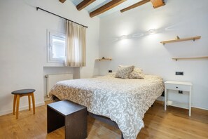 A bedroom with a bed, a bedside table, a small stool, a window with curtains, and wooden beams on the ceiling.