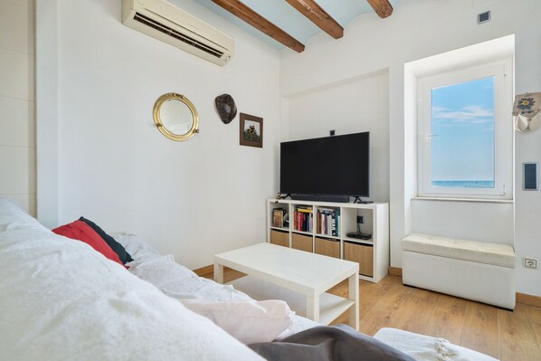 A living room with a white sofa, a white coffee table, a flat-screen TV on a white stand, a bookshelf with books, and a window with a view.