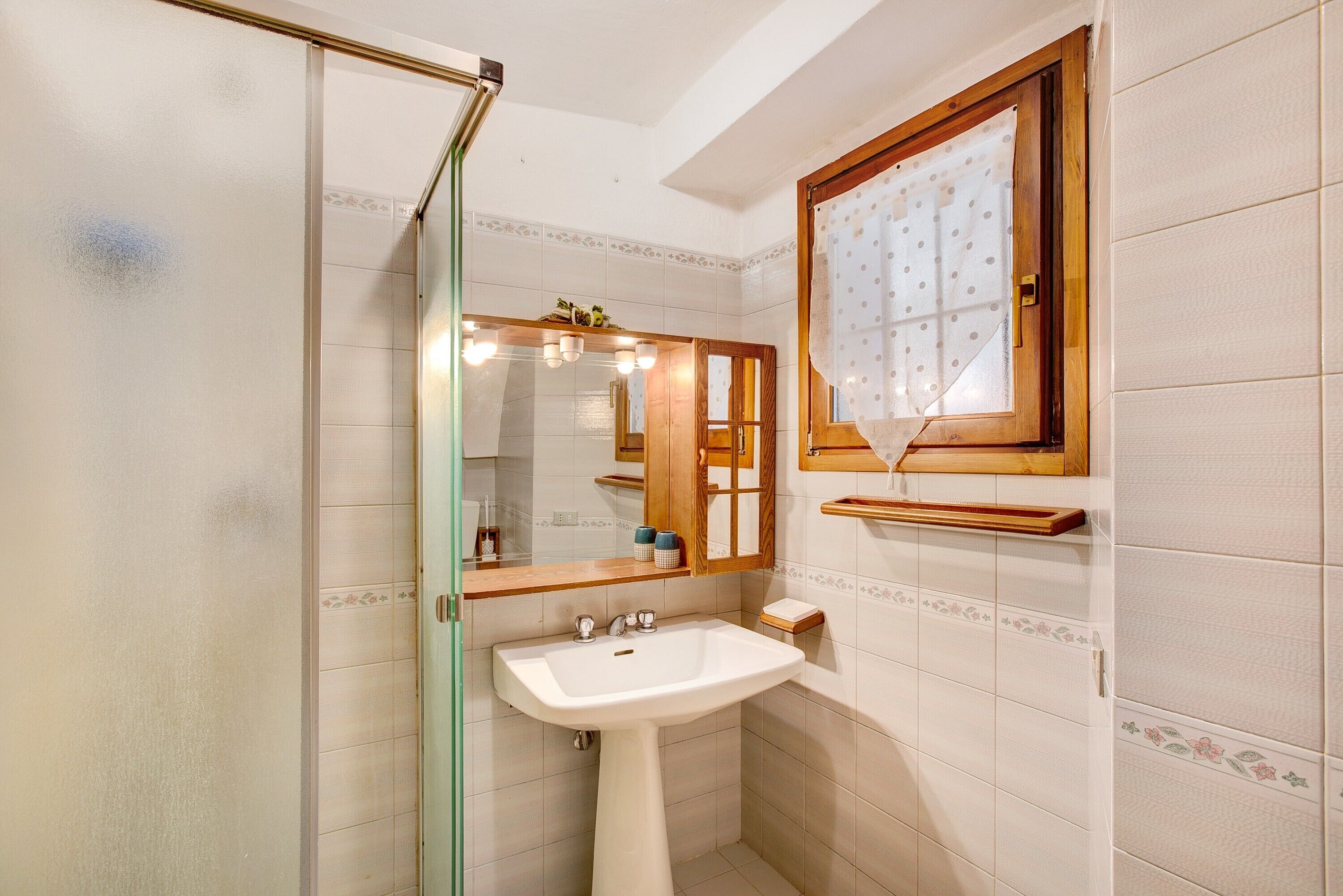 A bathroom with a glass shower enclosure, a pedestal sink, a mirror with a wooden frame, and a window with a polka dot curtain.