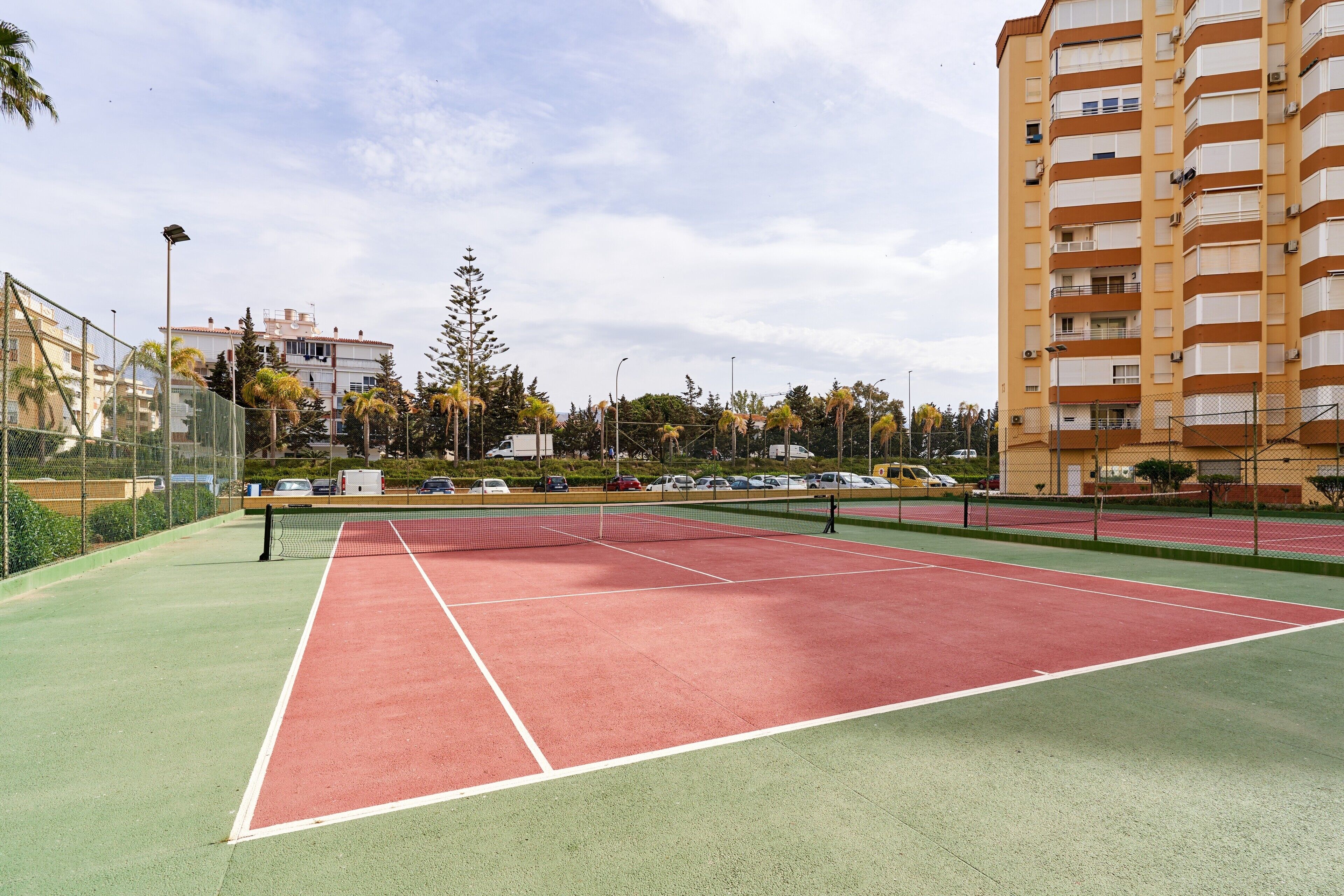 A tennis court with a net, surrounded by a fence, with buildings and parked cars in the background.