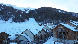 Un village de montagne enneigé, avec des bâtiments en bois, une piste de ski en arrière-plan et une route qui traverse le village.