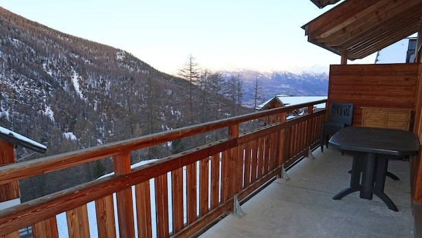 Un balcon doté d’une balustrade en bois, d’une table et de chaises. Il offre une vue sur un paysage montagneux enneigé.