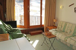 A cozy room with a wooden floor, a green sofa, a small table with a basket, a TV, and a window with a view of a snowy landscape. - Grand Studio 4 Personnes (LES ORRES 1650)