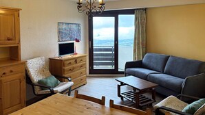 A living room with a wooden table, a sofa, a chair, and a TV on a wooden cabinet. There is a balcony with a view. - 2 Pièces 6 Personnes Centre Commercial (LES ORRES 1650)