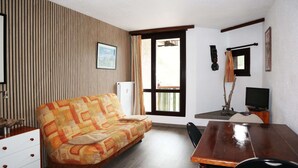 A room with a wooden slatted wall, a sofa, a wooden table, a chair, a television, and a window with curtains. - Studio Équipé Pour 4 Personnes (LES ORRES 1650)