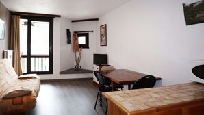 A room with a sofa, a wooden dining table, a TV, and a window with curtains. - Studio Équipé Pour 4 Personnes (LES ORRES 1650)