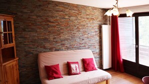 A room with a brick wall, a wooden cabinet, a sofa with red cushions, and a window with red curtains. - Grand Appartement 6 Personnes (LES ORRES 1650)