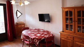 A dining area with a table covered in a patterned tablecloth, red chairs, a television mounted on the wall, and a wooden cabinet with glass doors. - Grand Appartement 6 Personnes (LES ORRES 1650)