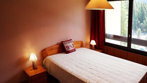 A neatly made bed with a white bedspread and a red pillow, two bedside lamps, a wooden nightstand, and a window with a view of trees. - Grand Appartement 6 Personnes (LES ORRES 1650)