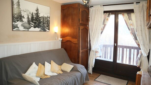 Una habitación acogedora con un sofá gris, un armario de madera, una ventana con cortinas y una terraza de madera afuera.