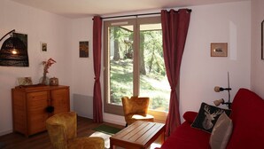 A cozy living room with a wooden dresser, a red sofa, a wooden table, and a chair. A large window with curtains offers a view of a forested area. - Appartement Spacieux, Idéal Pour une Famille (LES ORRES 1650)