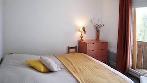A bedroom with a bed, a nightstand with a lamp, a window with curtains, and a view of greenery outside. - 2 Pièces 6 Personnes en Cœur de Station (LES ORRES 1650)