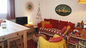 A cozy living room with a wooden dining table, a sofa with red and yellow cushions, a television, and a wall-mounted decoration. - 2 Pièces 6 Personnes en Cœur de Station (LES ORRES 1650)