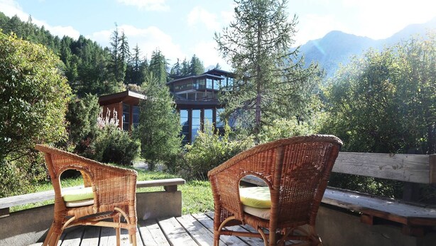 Deux fauteuils en osier, dotés de coussins, sont placés sur une terrasse en bois qui offre une vue sur un paysage montagneux, avec un bâtiment moderne en arrière-plan.