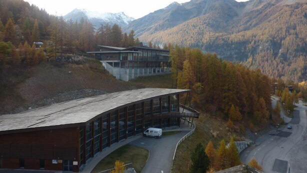 A modern building with a curved roof and large windows, surrounded by autumn-colored trees and mountains in the background. - 32 Orrianes des Forets (LES ORRES 1650)