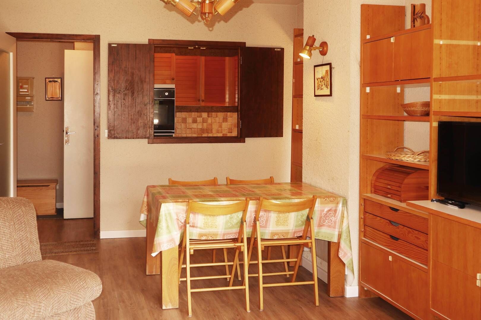A compact kitchen with wooden cabinets, a dining table with chairs, and a television on a wooden stand.