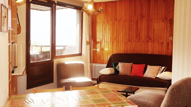 A cozy room with a wooden wall, a black sofa with patterned cushions, a dining table with chairs, and a window with curtains. - 834 Orrianes des Forets (LES ORRES 1650)