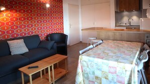 A compact living space with a sofa, a small table, a bed with a quilt, a kitchen area with cabinets, and a red patterned wall. - 2 Pièces 6 Personnes (LES ORRES 1650)