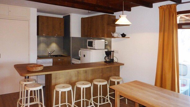 A compact kitchen with wooden cabinets, a microwave, a coffee maker, and a wooden table with bar stools. - 2 Pièces 6 Personnes (LES ORRES 1650)