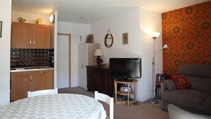 Een woonkamer met een televisie, een bank, een eettafel en een keukenhoek met kastjes en een betegelde achterwand.