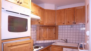 A kitchen with wooden cabinets, a white stove, a sink, and a tiled backsplash. - Appartement N°22 Eperviere (LES ORRES 1650)
