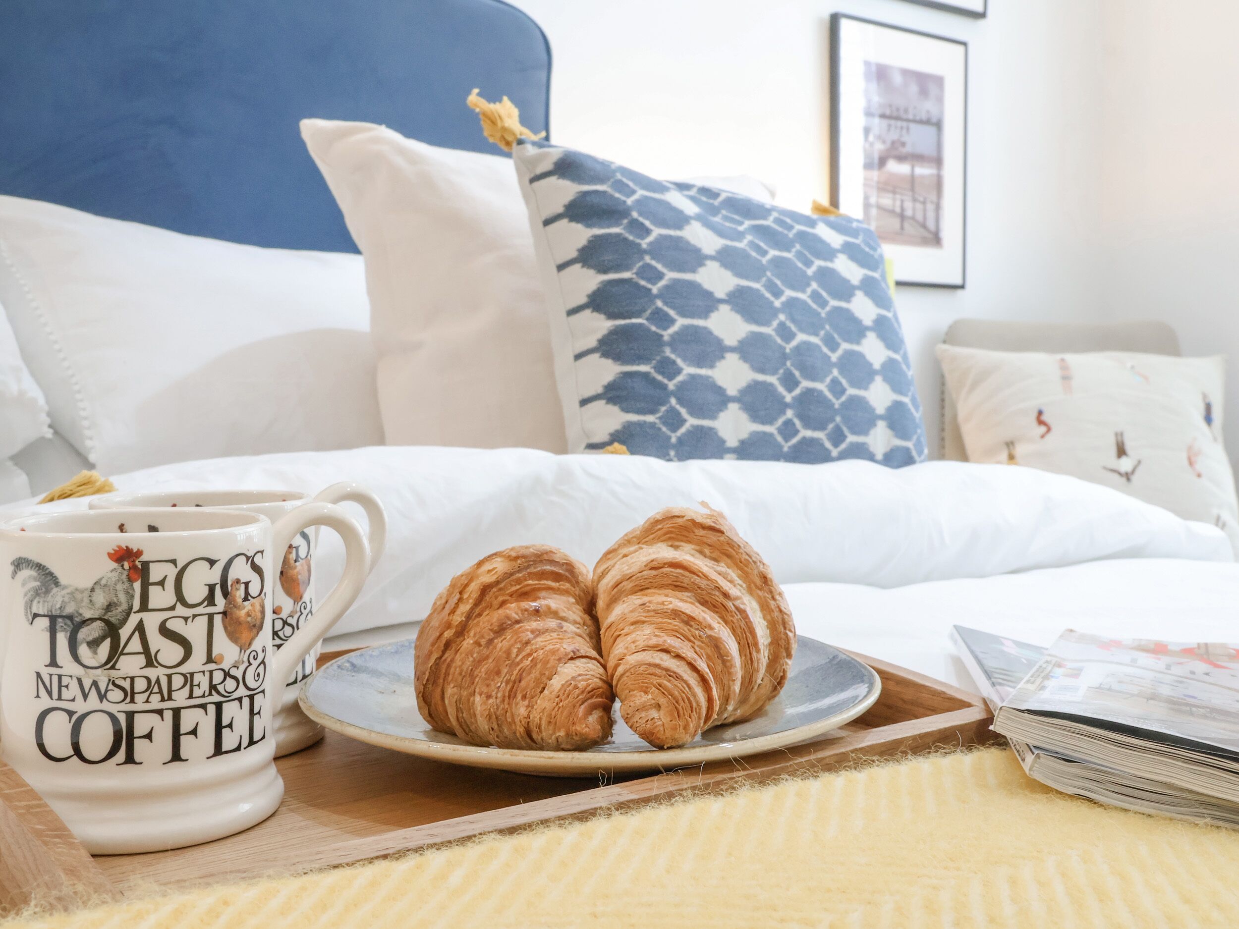 Una cama bien hecha con un cabecero azul, un plato con croissants y una taza con diseño de gallo.