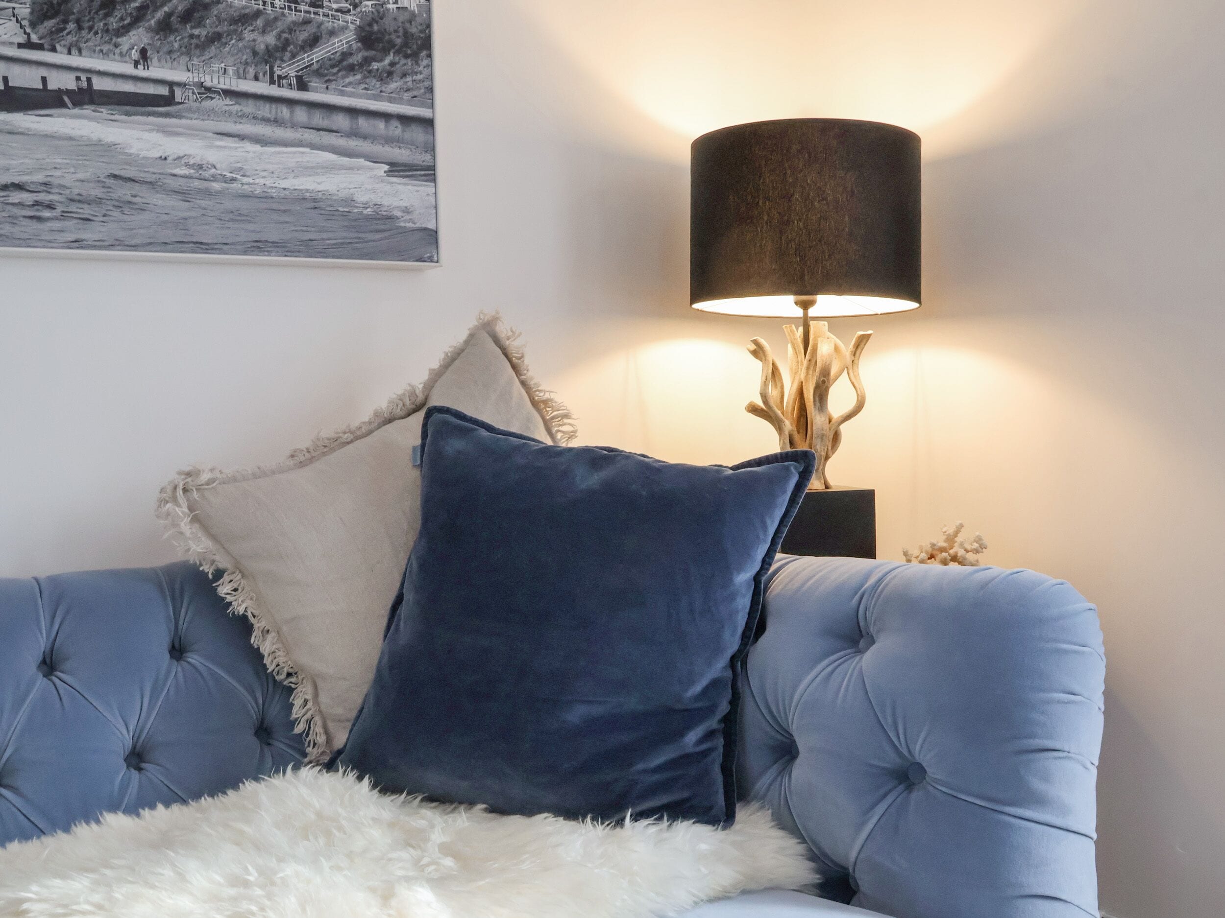 A room with a blue tufted sofa, a white fluffy throw, a dark blue pillow, and a lamp with a wooden base.